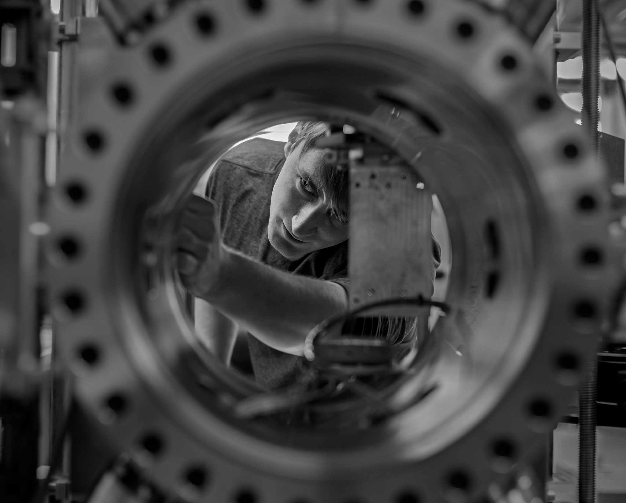 A black-and-white photo of a cryogenic engineer viewed through a circular opening of a large metallic chamber with evenly spaced bolts along its rim. Inside, a square component is mounted with wires connected to it. The man viewed through the porthole appears to be adjusting settings for the machine.