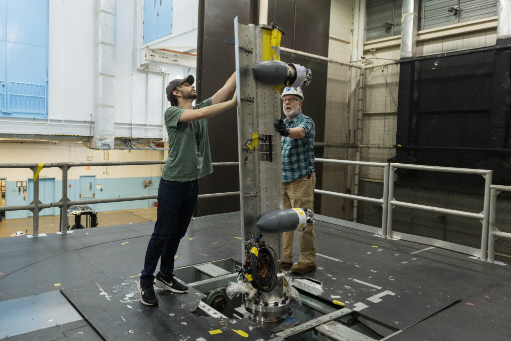 Two men are installing a vertical wing with mounted propellers inside a large wind tunnel facility.