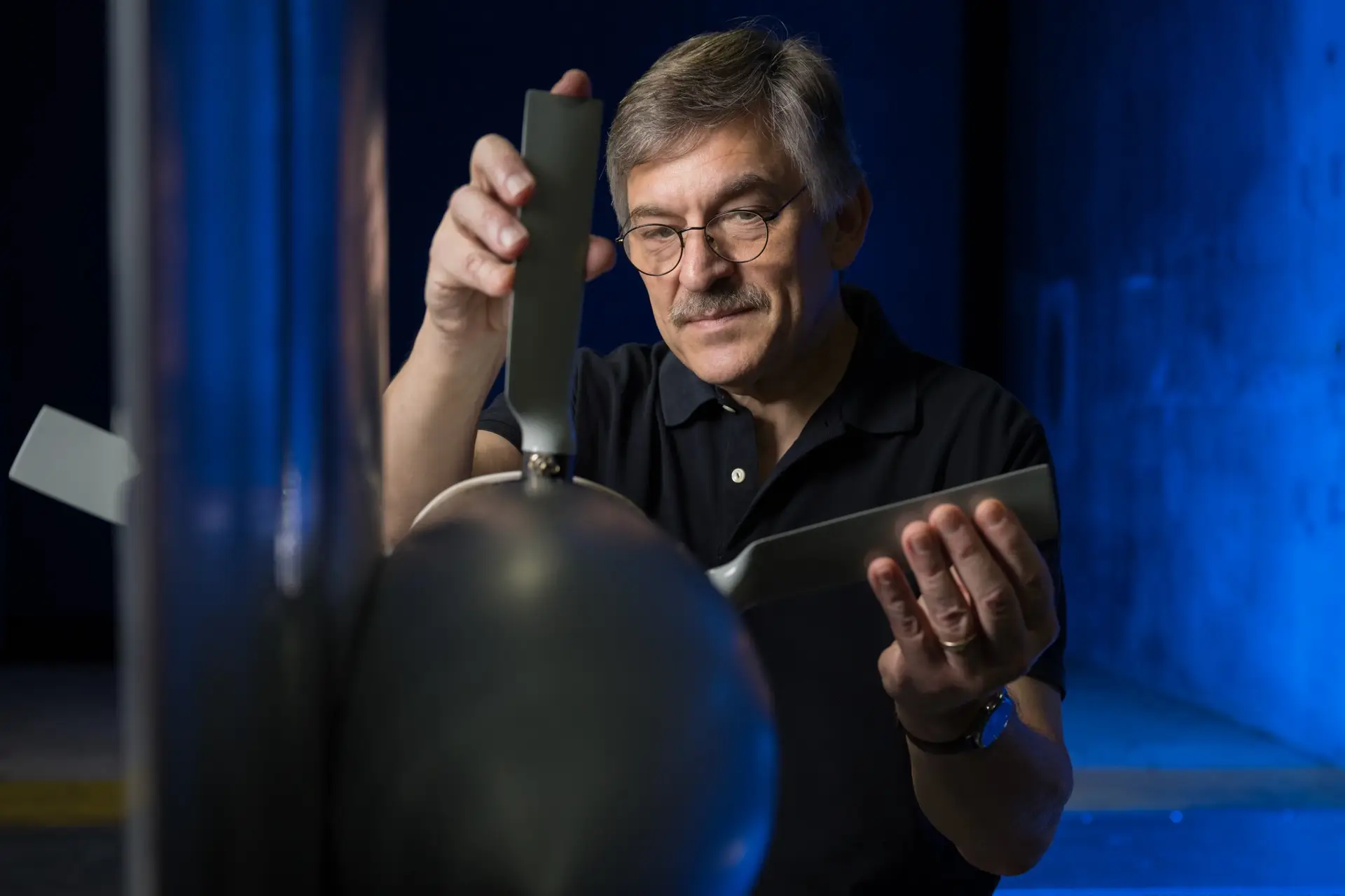 A man is adjusting a propeller blade attached to a test model, with a blue-lit background.