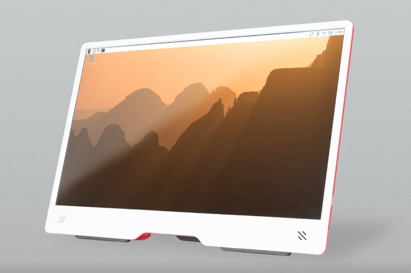 A Raspberry Pi Monitor displaying the new Raspberry Pi OS Trixie desktop wallpaper, which shows a golden sunrise over dramatic silhouetted mountain peaks