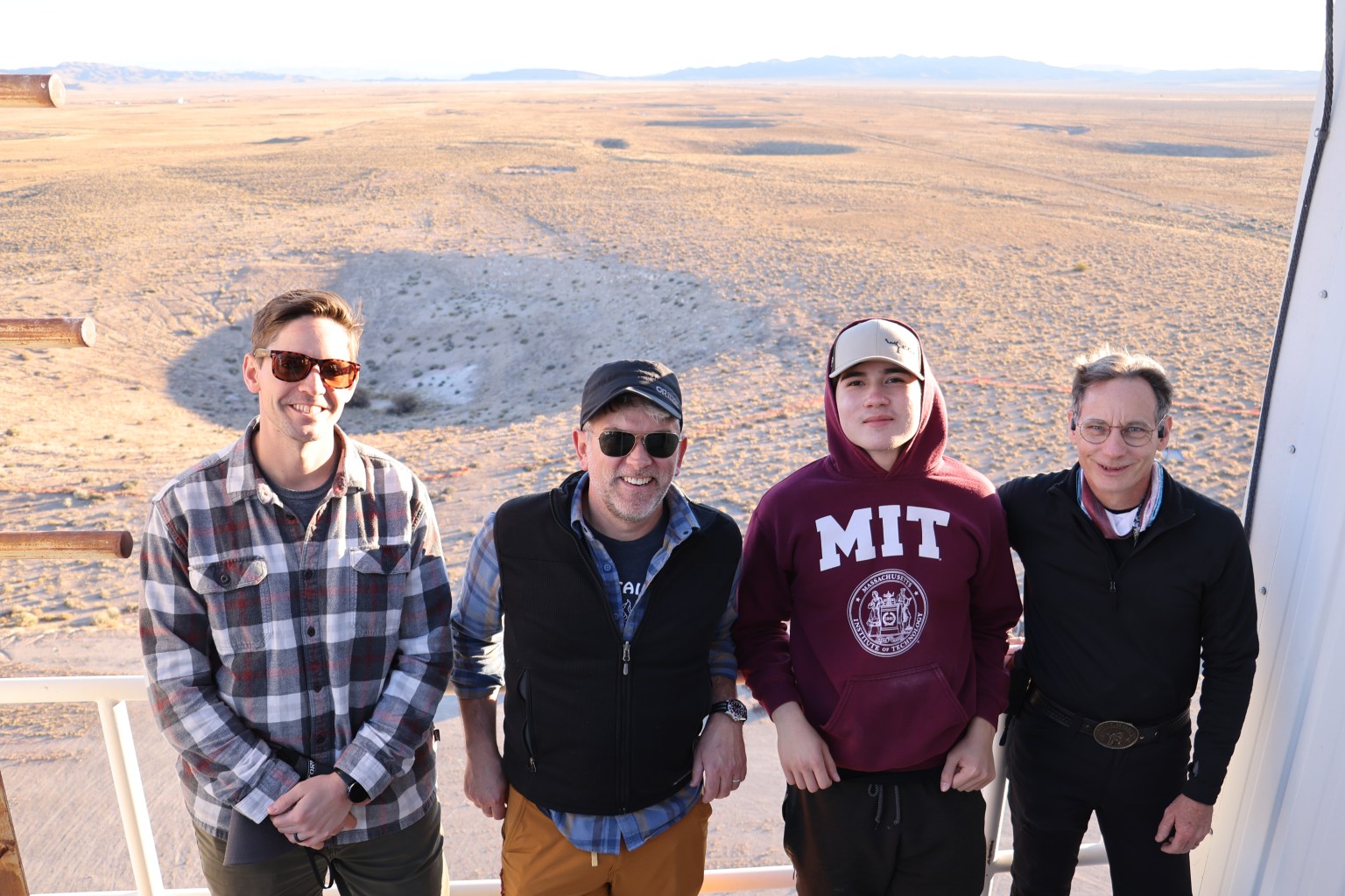 A group of people stand together at a test site in the desert, with wide open terrain and craters in the background.